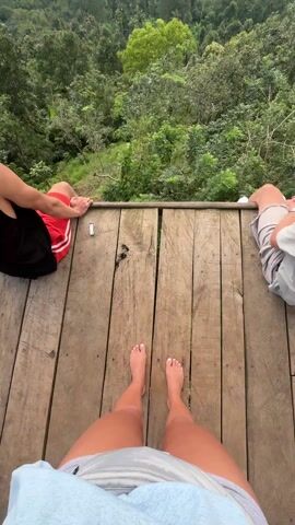 The video shows a view from a wooden platform overlooking a lush, green mountainous landscape. The sky above is overcast with dramatic clouds, and shafts of sunlight break through in places. At the beginning of the video, the camera focuses on the platform, showing bare feet and parts of the legs of three people sitting on the edge. One person wears red shorts, another gray, and the person holding the camera wears white shorts. As the camera pans up, one of the people, wearing a black tank top and red shorts, leans back to take in the view.