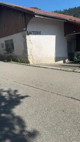 The video shows a motorcycle riding down a paved road in a village. The motorcycle is black with a red top case and is ridden by a person wearing a helmet and dark clothing. The road is lined with houses and trees. The sky is clear and blue. The motorcycle is moving away from the camera and appears to be going uphill. The word "LAITERIE" is visible on the side of one of the buildings.