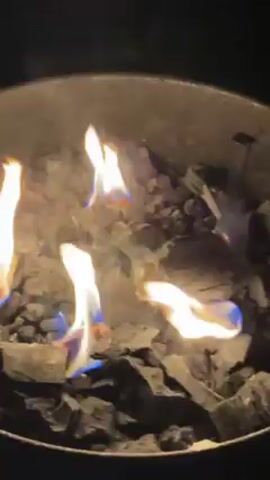 The video shows a metal fire pit filled with burning charcoal.  Bright, nearly transparent flames with blue edges flicker across the charcoal, illuminating it against the dark surroundings.  A single, slender object falls from above and briefly enters the frame.