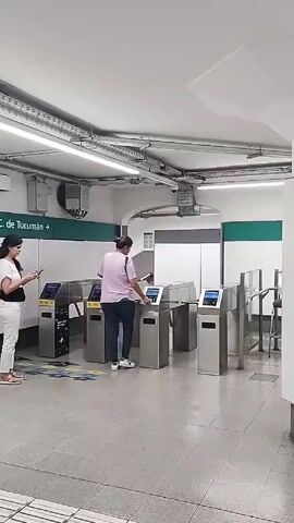 Two women are at the turnstiles of a subway station. The woman to the left, dressed in white pants and a beige shirt, is looking at her phone. The woman on the right, wearing jeans and a pink shirt, has just gone through the turnstile and is walking away. The turnstiles are made of silver metal with black accents. An arrow on the wall points to the right, indicating the direction of C. De Tucumán. The station has white walls, gray floors, and visible wiring on the ceiling.