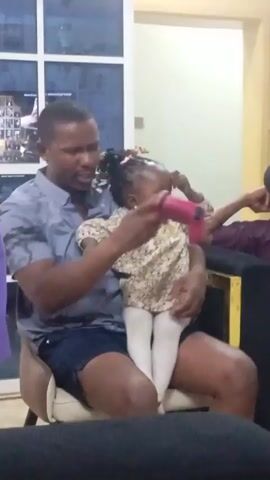 A man is sitting in a chair holding a young girl on his lap. The man is wearing a grey and blue camouflage-patterned shirt and dark blue shorts. The girl is wearing a light-colored, floral-patterned dress and white tights. She holds a small pink object in her hands. Another girl in a white shirt is visible to the left of the man. An unidentified person is seated to the right of the man. The man is talking, and the girl is looking down at the object in her hands. The scene appears to be indoors, with a window behind the man. A framed photo hangs on the wall behind the window. A dark couch and a yellow object are also visible in the background.