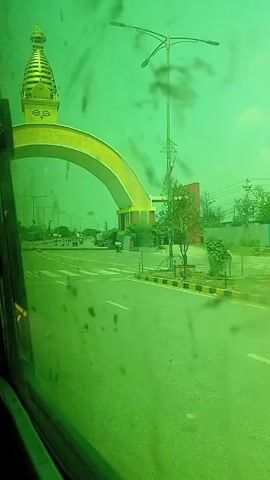 The view from a vehicle passing under a large golden archway with a stupa-like structure on top is captured in this short video. The footage is tinted green, likely due to the vehicle's window tint. 

A road stretches into the distance, with vehicles like motorcycles and what appears to be an auto-rickshaw visible. A pedestrian on a bicycle is also visible on the road. The archway leads to a building or entrance with a small garden area. Utility poles are scattered along the roadside.
