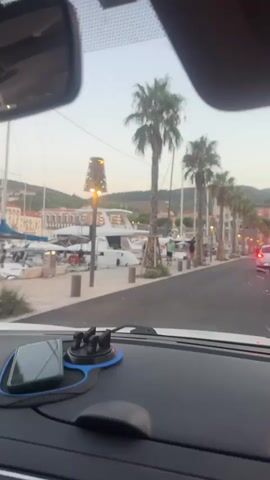 This is a first-person perspective video taken from inside a car. The car is driving down a paved road lined with palm trees on the left side. Adjacent to the palm trees is a marina with several yachts docked. Across the road on the right, there are buildings and more palm trees. The sky is overcast, suggesting it is either dawn or dusk. There are a few people walking along the sidewalk near the marina and a few cars driving on the road. A phone is mounted on the dashboard in front of the driver.