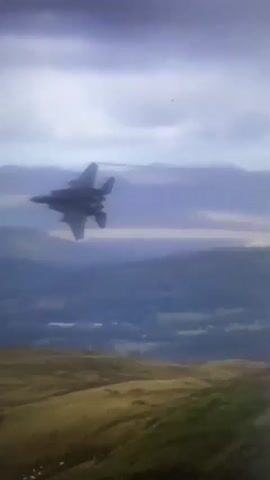 A fighter jet performs a maneuver over a mountainous landscape, leaving a contrail in its wake. The terrain below consists of rolling hills and valleys, covered in a mix of vegetation and bare rock. The sky is overcast with some visible clouds. The camera angle shifts, providing different perspectives of the jet and its surroundings.