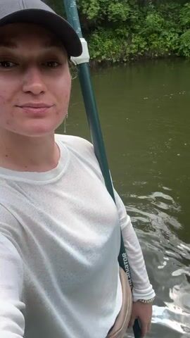 A person with a dark baseball cap, light long-sleeved shirt, and light fanny pack stands in the foreground, filming themselves with a camera that captures their face and upper body. The background shows a river with green water, bordered by a green forested bank. Two individuals are visible in the distance, each standing on a paddleboard. One person is positioned further down the river, while the other is closer to the bank. A paddleboard and a pair of sandals are visible in the bottom right corner of the frame. The video depicts a calm outdoor scene.