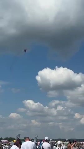 A red jet fighter performs an aerial maneuver against a blue sky dotted with white clouds. Below, a crowd of people watch the display. The jet is seen in the upper center of the frame, leaving a faint smoke trail. Some of the spectators are holding up phones to record the event. The overall scene suggests an airshow.