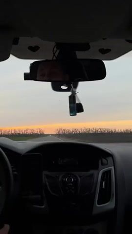 The video captures a first-person perspective from inside a car driving on a straight road. The driver's face is partially visible in the rearview mirror, showing their eyes and forehead as they look ahead.

The car's interior is dark, with the dashboard, steering wheel, and infotainment system visible. A smartphone is mounted on the left side of the dashboard. A blue and black air freshener hangs from the rearview mirror. The car's headliner above the mirror is decorated with a pattern of small black hearts.

Outside the car, the sky displays warm orange and pink hues along the horizon, indicating either sunrise or sunset. A line of bare trees is silhouetted against the colorful sky in the distance. The road appears to be a clear, two-lane highway extending into the horizon.

On the car's infotainment display, some numbers like "10:56" are faintly visible, along with other unreadable text, likely relating to the radio or time. An orange indicator light is also illuminated on the dashboard. Throughout the video, music with a noticeable beat is playing in the background. The scene depicts a tranquil drive, likely at dusk, in the Starominskaya, Russia area.