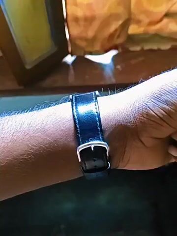 A black leather banded watch with a silver buckle is fastened around a person's wrist. The watch has a black face with silver hands and markers, and a silver casing with two buttons on the right side. The background is out of focus, showing what appears to be an orange curtain or fabric. The camera slowly zooms in on the watch.