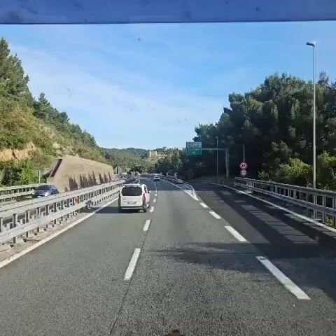 The video shows a view from a moving vehicle on a highway. The highway has multiple lanes separated by white lines and is flanked by concrete barriers and guardrails. Other vehicles are visible in the distance.  A green highway sign indicating the direction to Andorra is present. The camera zooms in on a medieval-style building complex situated on a hilltop, surrounded by trees and foliage. The sky is clear and blue.
