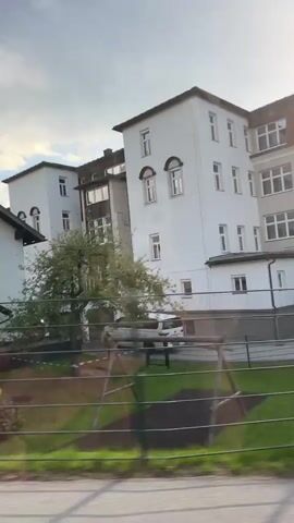 This video appears to be a recording from a moving vehicle, likely a car or bus, traveling through a residential area. The camera captures a side view of buildings, including what looks like a school or institutional building with multiple stories and many windows. In the foreground, there's a glimpse of a playground with a yellow slide. The scene is bordered by a fence and trees, suggesting a suburban or semi-rural setting.

The time of day appears to be late afternoon or early evening, given the soft, warm light and the angle of the sun. The sky is partly cloudy, with bright sunlight breaking through, creating a somewhat dramatic lighting effect. The overall mood is peaceful and ordinary, depicting everyday life.

The location context points to Sankt Johann im Pongau, Austria. While no specific text is clearly visible on the buildings or signs, the architecture and the presence of the mountains in the background are consistent with a European setting. The footage is shaky, as expected from a moving vehicle, and the focus shifts as the camera captures different elements of the passing scenery.