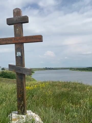 The video begins with a close-up of a wooden cross standing in tall grass. The cross is weathered and has some text and a small icon on it. The camera then pans to the right, revealing a wide river flowing through a landscape. The river is calm with a slight ripple. On the far bank, there is a dense forest. The sky is cloudy with patches of blue visible. The grass in the foreground is green and yellow, with some wildflowers scattered throughout. The video continues to pan, showing more of the river and the forest. The overall impression is one of a peaceful, natural setting.