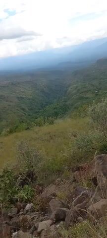 The camera pans across a green, hilly landscape under a cloudy sky. The viewpoint is elevated, looking down into a valley filled with trees and vegetation. The foreground features dry grass and rocks.