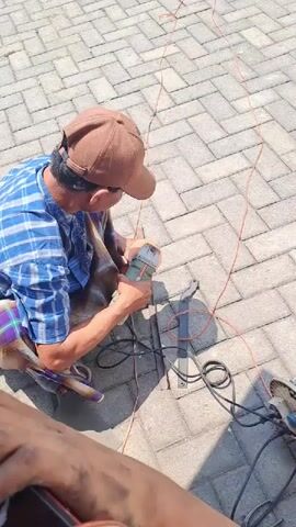 A person wearing a blue and white checkered shirt, a brown baseball cap, and a sarong is kneeling on a brick-paved surface. The person is holding a grinder tool and appears to be working on something. There are several red wires and black power cords on the ground, as well as another grinder tool. The scene is outdoors, with sunlight casting shadows.