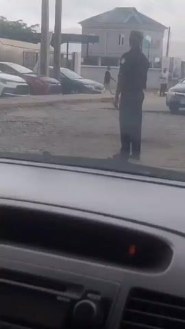 The video shows a street scene in Jibowu, Nigeria. A person in a dark uniform, possibly a police officer, stands in the middle of the road, seemingly directing traffic. Several cars are visible, including a black sedan and a red car, parked or moving. In the background, there are buildings and utility poles. The sky is overcast, suggesting a cloudy day. The overall impression is of a busy urban environment. There is no visible text in the video.