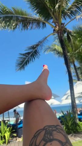 The video shows a person lying down at what appears to be a beach resort. Their leg is raised with their foot resting on their thigh. The toes are painted with red nail polish. A tattoo is visible on their thigh. In the background, palm trees sway under a clear blue sky. There are other people relaxing on lounge chairs with white umbrellas nearby, and the ocean is visible in the distance.