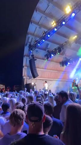 The video shows a concert at night. In the foreground, a crowd of people is visible, with many holding up their phones to record. The crowd is illuminated by blue and green stage lights. In the background, a performer with a green hat is singing into a microphone on stage. Behind the performer, other band members are visible, as is a stage structure with lights and speakers. The overall atmosphere appears energetic and celebratory.