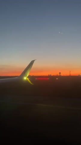 A low-angle shot from inside an airplane shows a Ryanair wing with its yellow accents brightly lit against a stunning sunrise or sunset. The sky transitions from a deep orange and pink horizon to a soft blue overhead. The aircraft appears to be stationary on a dark runway, with red lights visible in the distance, likely marking the runway edge or approach lights. Further in the background, dimly lit structures or a distant cityscape can be seen. The wing's anti-collision lights are visible, and the sound of an aircraft engine is audible. The scene depicts an early morning or late evening at an airport in Store Magleby, Denmark.