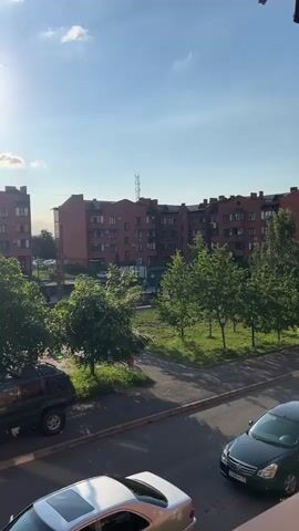 The video shows a residential area with buildings, trees, and cars. The sun is shining brightly in a clear blue sky with a few clouds. In the foreground, a silver car with a sunroof is parked. Further back, a dark green SUV is parked. Beyond the cars, there are trees and a playground. In the background, red brick apartment buildings are visible. A person in a red outfit is briefly seen walking on a path between the trees. The scene appears to be a sunny day in a suburban or urban environment.