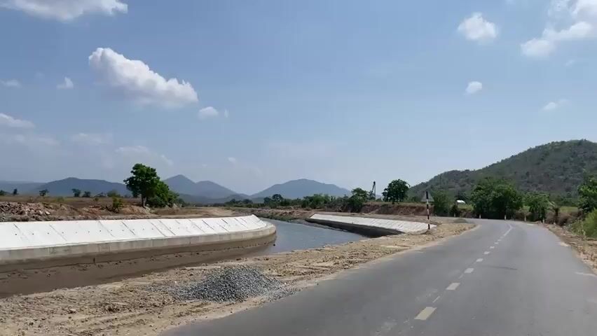 The camera pans across a rural landscape featuring a concrete-lined irrigation canal running alongside a paved road. The canal contains water and has banks lined with reddish-brown soil. A road sign is visible near the canal. The road curves gently to the right. In the background, there are hills and mountains under a blue sky with scattered white clouds. Trees are sparsely distributed across the landscape.