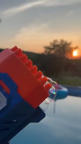 A bubble machine is pointed towards the right side of the frame and is producing several bubbles. The bubble machine is blue and orange with multiple orange nozzles. Behind the bubble machine, the sun is setting in the background, casting an orange glow on the sky. There are trees and a hill in the distance. In the foreground, there is a blue swimming pool.