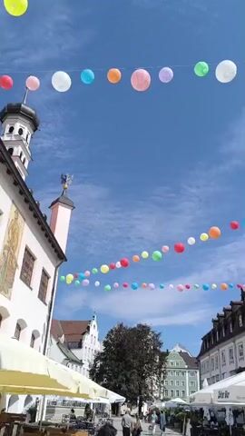 The video shows a street scene with colorful paper lanterns strung across the sky. The lanterns are of various colors including red, orange, yellow, green, blue, and pink. They are suspended at different heights, creating a festive atmosphere. Below the lanterns, there are buildings with traditional European architecture. Some buildings have awnings and outdoor seating areas, suggesting the presence of cafes or restaurants. In the background, there are people walking on the street. The sky is blue with some scattered clouds, indicating a clear day. The camera pans and zooms, capturing different perspectives of the street and the lanterns.