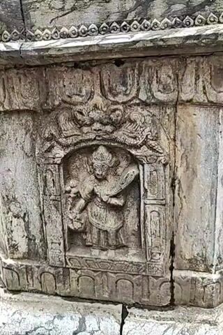 A stone structure with intricate carvings is filmed from various angles. The structure appears to be part of a larger monument or temple, with additional decorative elements and inscriptions visible at the top. In the background, glimpses of other buildings and greenery suggest an outdoor setting, possibly a courtyard or a public space. There are no people visible in the video, and the focus remains solely on the stone artifact.