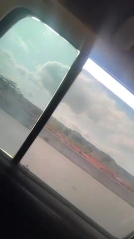 A handheld camera inside a car captures a view of a highway and hillside through a partially open tinted window. The sky is partly cloudy. Music plays.