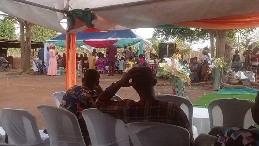 The video captures an outdoor gathering under a tent. The setting appears to be a rural area, with trees surrounding a dirt ground. Numerous individuals are present, some seated and others standing around a central area decorated with colorful fabric. In the foreground, people sit in white chairs, their backs to the camera. The crowd is dressed in a variety of colorful attires. A few floral arrangements are visible near the front. The overall atmosphere suggests a celebratory or formal event.