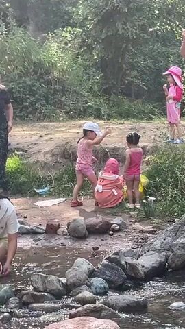 A group of people are gathered near a rocky stream. Two young girls in pink swimwear and hats are playing near the water's edge. One girl is crouching, looking at something on the ground, while the other stands beside her. A woman in a black shirt and patterned pants stands to their left, watching them. To the right, a boy wearing blue shorts and a pink hat is holding something to his mouth. Another young girl in pink is standing near him. The area is surrounded by lush green trees and vegetation, with rocks and dirt visible on the bank of the stream.