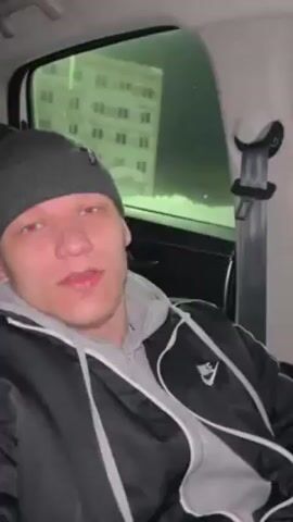A light-skinned man is in the back seat of a car at night, looking directly at the camera while speaking. He is wearing a grey beanie hat, a grey hooded sweatshirt, and a black puffy jacket with white trim. The background visible through the car window suggests an urban setting, with a lit apartment building partially visible against the dark night sky. The camera is steady.