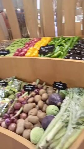 The video displays a brightly lit, modern grocery store setting featuring a large, multi-tiered wooden stand showcasing fresh produce. The display is curved and stacked, presenting an array of colorful fruits and vegetables.

On the upper tiers, various bell peppers (yellow, red), green beans, and beetroots are neatly arranged. The lower tiers feature a diverse selection, including cherry tomatoes (some packaged in plastic containers, others loose), bundles of leafy greens (many individually wrapped in plastic), onions, potatoes, green and purple cabbages, and what appear to be asparagus. Some items are displayed loose, while others are pre-packaged in clear plastic or mesh bags.

Small black labels with white text are placed in front of different produce items, likely indicating names or prices, though the text itself is not legible. The background consists of modern light-colored wooden panels, and a glimpse of a purple screen is visible on the left side of the frame at one point. The overall impression is that of a well-organized and aesthetically pleasing produce section within a supermarket. The location is specified as Al Machouar – Stinia, Morocco.