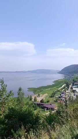 The video shows a wide, calm river with a distant hazy shoreline. On the near side of the river, a sandy beach with several people and tents is visible. Adjacent to the beach, dense green reeds and grass grow along the riverbank. Further inland, a small village with houses and a road winds its way along the slope. The sky is bright blue with a few scattered white clouds. The overall impression is a serene summer day at a riverside location.