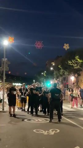 A group of people are playing drums in the street at night. They are all wearing black clothing and are facing forward. There are spectators on the side of the street watching them. There are also flower-shaped lights hanging from the trees. The street is paved and has white lines painted on it, including a bicycle lane. The sky is dark blue.