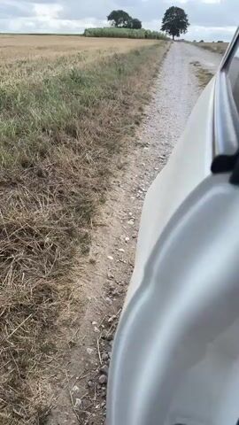 The video shows a perspective from inside a moving white car driving on a gravel road through a rural landscape. The road is lined with dry grass and fields. In the distance, there are trees and more fields, with a blue sky and white clouds overhead. The car's side mirror and part of its interior are visible in the frame. The camera pans and tilts as the car moves, capturing the scenery.
