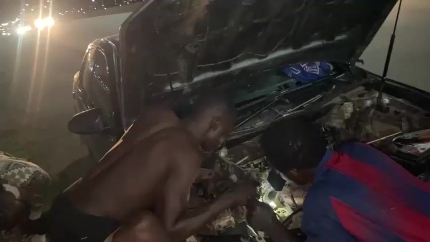 At night, three men are working on the engine of a car with its hood open. One man, shirtless, is leaning into the engine bay. Another man, wearing a blue and red striped shirt, is also looking into the engine bay, holding a tool. A third person, partially visible, is also near the engine. The car appears to be on the side of a road, with streetlights and traffic in the background. A bus with "CNG BUS" written on the back drives past the scene.