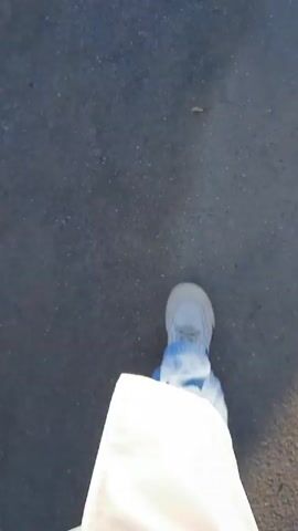 The video shows a first-person view of someone walking on a paved surface. The pavement appears to be dark gray and has some ice or snow on the edges. The person is wearing white clothing and light-colored shoes with blue accents. The camera is mostly pointed downwards, focusing on the person's feet and the ground.