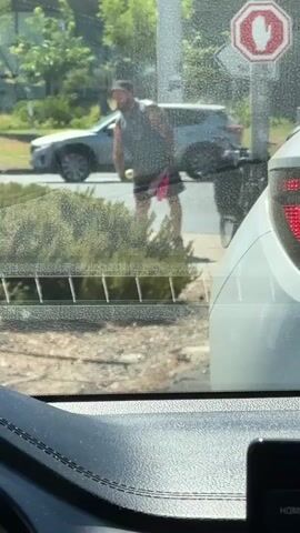 A person wearing a grey tank top, dark shorts, and a hat is standing by a road, holding a pink object. They are near a street sign and some bushes. The scene appears to be outdoors in daylight. The video is shot from inside a vehicle, as indicated by the interior car elements visible in the foreground.