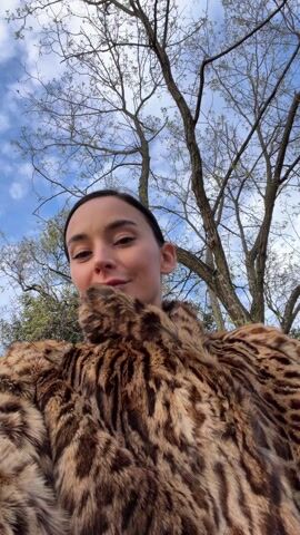 A woman wearing a fur coat is sitting on a swing in a park. The swing set is red. There is grass in the background. The woman is smiling. The camera is at eye level, pointing up at the woman. The background features trees and a blue sky with some clouds. The camera moves as the swing moves.