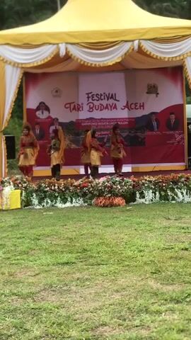 A group of five young female dancers in traditional attire are performing on a stage. The dancers are wearing red tops, yellow skirts, and orange headscarves, and they are holding orange cloths above their heads. They are arranged in a line across the stage and appear to be moving in unison. The stage is decorated with flowers and has a backdrop that reads "Festival Tari Budaya Aceh," which translates to "Aceh Cultural Dance Festival." The backdrop also features images of men in suits, presumably dignitaries. The stage is set up outdoors in a grassy area, with a yellow tent covering the top. The lighting suggests it is daytime. The dancers' movements are graceful and synchronized.