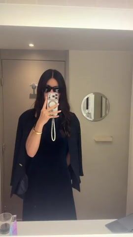 A woman stands in a bathroom, taking a mirror selfie with her phone. She has long, dark hair and wears sunglasses, a navy dress, and a black blazer draped over her shoulders. She is also wearing a bracelet and a pearl necklace. The bathroom has a tiled wall, a vanity with a circular mirror, and a stainless steel door. There are a few items on the counter, including a glass and a laptop. The room is well-lit, and the woman is the main focus of the shot. She is posing for the photo, raising her fist slightly.