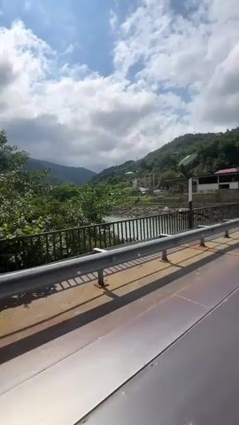The video shows a view from a bridge overlooking a river in a mountainous region. The river is a turquoise color and appears to be flowing. The banks of the river are lined with lush green trees and vegetation. In the distance, rolling green hills and mountains rise up, partially obscured by clouds. The sky is blue with scattered white clouds, and the sun is shining. There is a metal railing along the edge of the bridge, and a lamppost with a solar panel is visible. A few buildings can be seen on the hillside. The perspective suggests the video was taken from a moving vehicle on the bridge, as indicated by the subtle vibration and the slight shift in the background scenery.