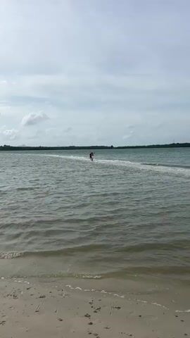 A person on a jet ski is moving across a calm body of water, approaching a narrow sandbar that extends into the water. The sky is overcast with scattered clouds. The water is a murky green color, with small waves lapping at a sandy shore in the foreground. The sandbar is light in color and appears to be above the water level. The person on the jet ski is wearing dark clothing and is seated.