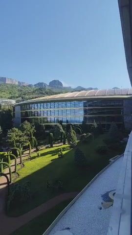 The video pans slowly to the left from a view of a multistory building with a curved glass facade, toward a landscape with mountains in the background. The building is surrounded by a manicured lawn with arched hedges and a paved path. The landscape beyond the building features lush green trees and other structures, possibly houses. The sky is clear and blue. The lighting suggests it is daytime. At the bottom of the frame, there is a white-tiled area, possibly a balcony. An audible wheezing cough is also present.