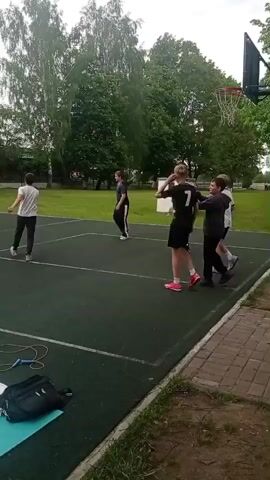 The video shows a group of adolescent boys on an outdoor basketball court. The court is green with white lines and a basketball hoop at the end. One boy dribbles a basketball, while the other boys are standing around. Behind the court is a grassy lawn with trees. Off to the side of the court is a small bag and jump rope.