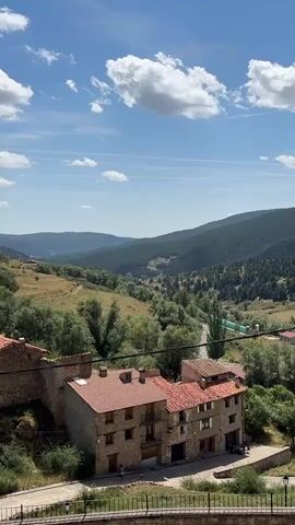A landscape video displays a rural village with red-roofed houses in the foreground. The buildings are made of stone and appear to be several stories high with small windows. Beyond the village, rolling hills covered in green trees and vegetation recede into the distance. A winding road is visible in the valley. The sky is blue with scattered white clouds, suggesting a sunny day. The video is shot from an elevated perspective, looking down on the scene. There are no people or animals visible.