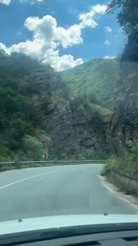 The video shows a first-person perspective from a vehicle driving on a winding road through a mountainous, forested area. The road is paved with white lines indicating lanes. On the left side of the road, there are steep, rocky cliffs covered with greenery, and a guardrail. On the right side, the road is bordered by a concrete barrier and more vegetation. In the distance, a white van is visible driving in the same direction. The sky is blue with scattered white clouds. The sun is shining, casting shadows. The overall impression is a scenic drive through a natural landscape.