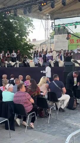 The video shows an outdoor performance on a stage in front of an ornate building. A group of performers, some dressed in traditional folk costumes, are on the stage, with a speaker addressing the audience. Several people are seated in the audience, facing the stage. A cameraman is filming the event. The building behind the stage appears to be a historic structure with balconies and decorative architecture. The sky is visible and appears to be daytime.