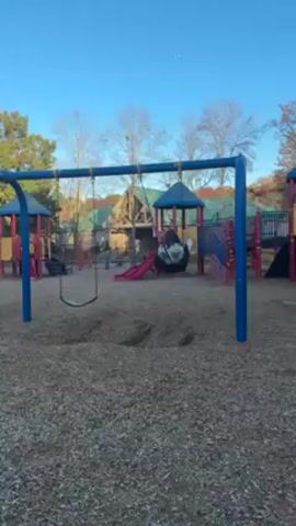 A woman is swinging on a swing set at an outdoor playground. The playground is covered in woodchips. In the background, there is a colorful playground structure with a slide. Other playground equipment is visible, as well as some trees without leaves. The woman is wearing a colorful tie-dye shirt, black pants, white shoes, and a black jacket that is not zipped up. She is facing away from the camera and swinging back and forth. The sky is blue with some clouds.