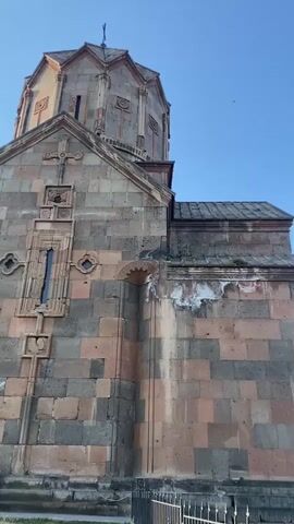 This media file features a close-up view of an old stone church, likely religious architecture, with a clear blue sky above. The church is constructed from large, rough-hewn stones in varying shades of pink, brown, and grey, displaying intricate carvings and architectural details. A cross is visible at the apex of the structure. The camera then pans to reveal a breathtaking landscape.

The scene transitions to a panoramic view of a deep canyon under a bright blue sky dotted with fluffy white clouds. The canyon walls are rugged and reddish-brown, with sparse vegetation clinging to the slopes. Across the canyon, a vast, rolling green landscape stretches towards a majestic mountain range in the distance. The sunlight suggests it is daytime, likely late afternoon given the warm lighting on the landscape. The overall atmosphere is serene and awe-inspiring, highlighting the natural beauty of the setting. The church appears to be perched on the edge of the canyon, offering stunning views. No people are clearly visible in the initial church shot, but in the canyon view, a person can be seen sitting on the edge of the canyon on the left side of the frame. No text is visible. The location is identified as Ohanavan, Armenia.