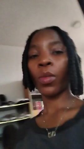 The video begins with a close-up of a woman with braided hair, looking directly at the camera. She is wearing a dark t-shirt and a necklace with a V-shaped pendant. The background is slightly blurred, but suggests an indoor setting with shelves filled with items that appear to be books or boxes. As the video progresses, the camera moves slightly, and the woman smiles. The lighting appears to be natural, possibly from a window. The woman speaks, but her words are not transcribed. The overall scene is casual and personal, likely a selfie-style recording.